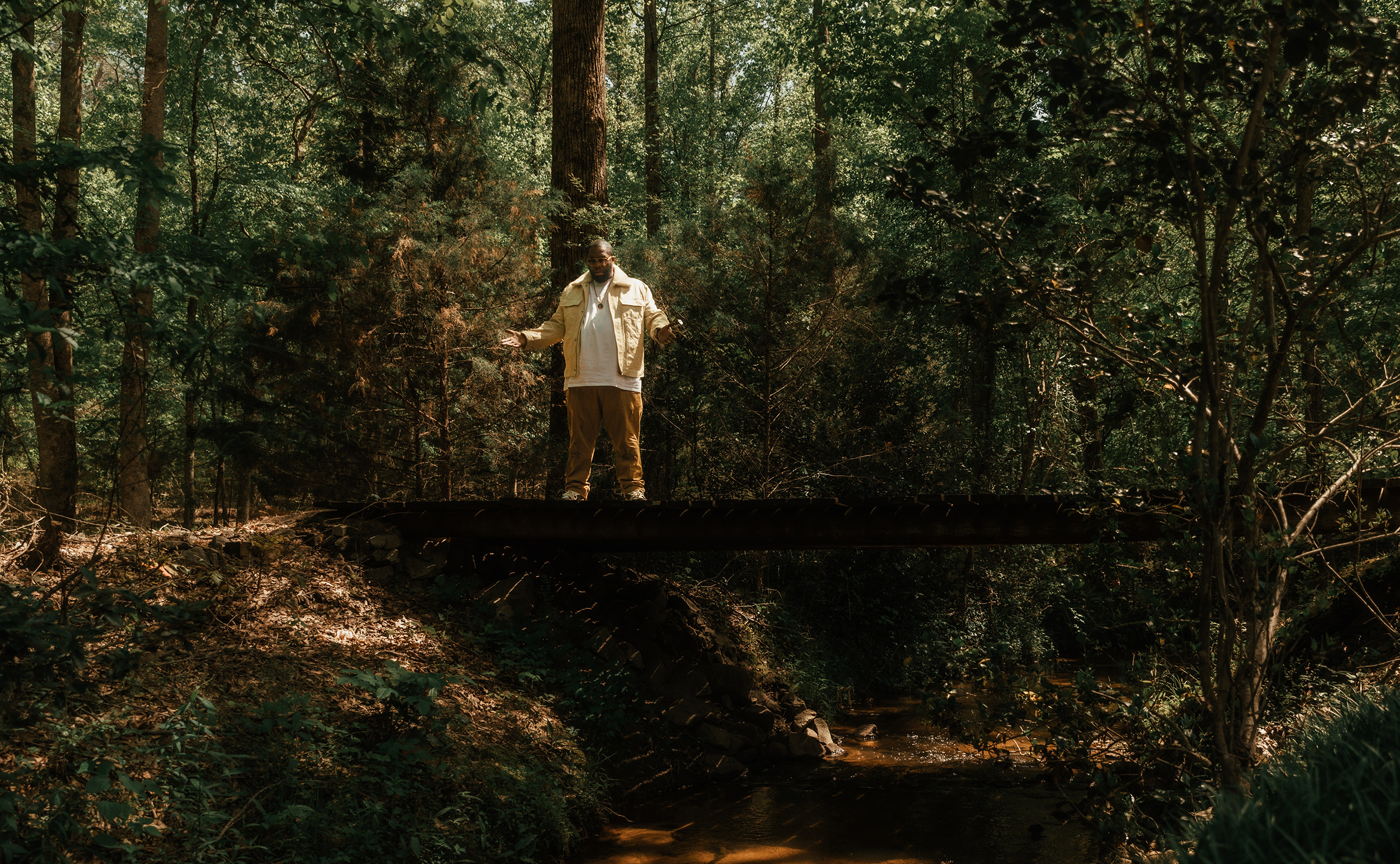 Killer Mike stands on a wooden bridge over a stream in the woods