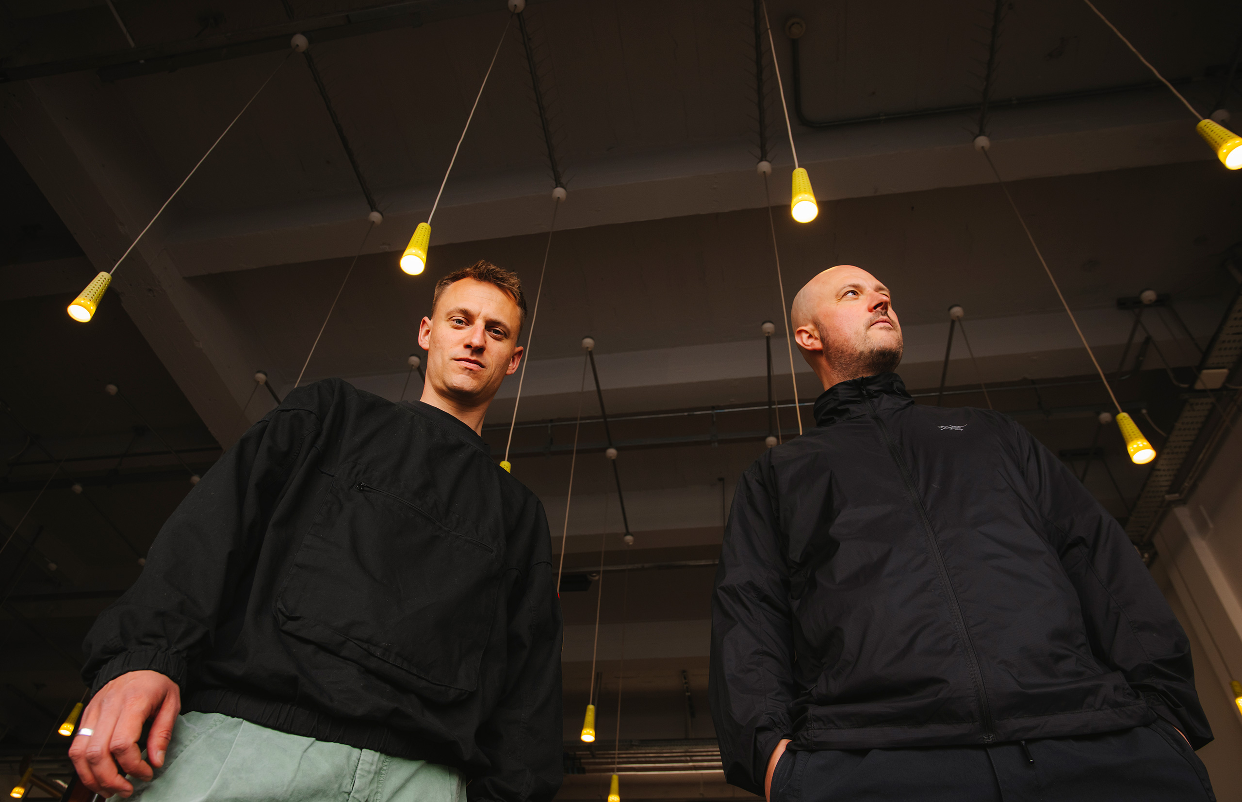 The two members of Overmono looking down at the camera with hanging lightbulbs above their heads