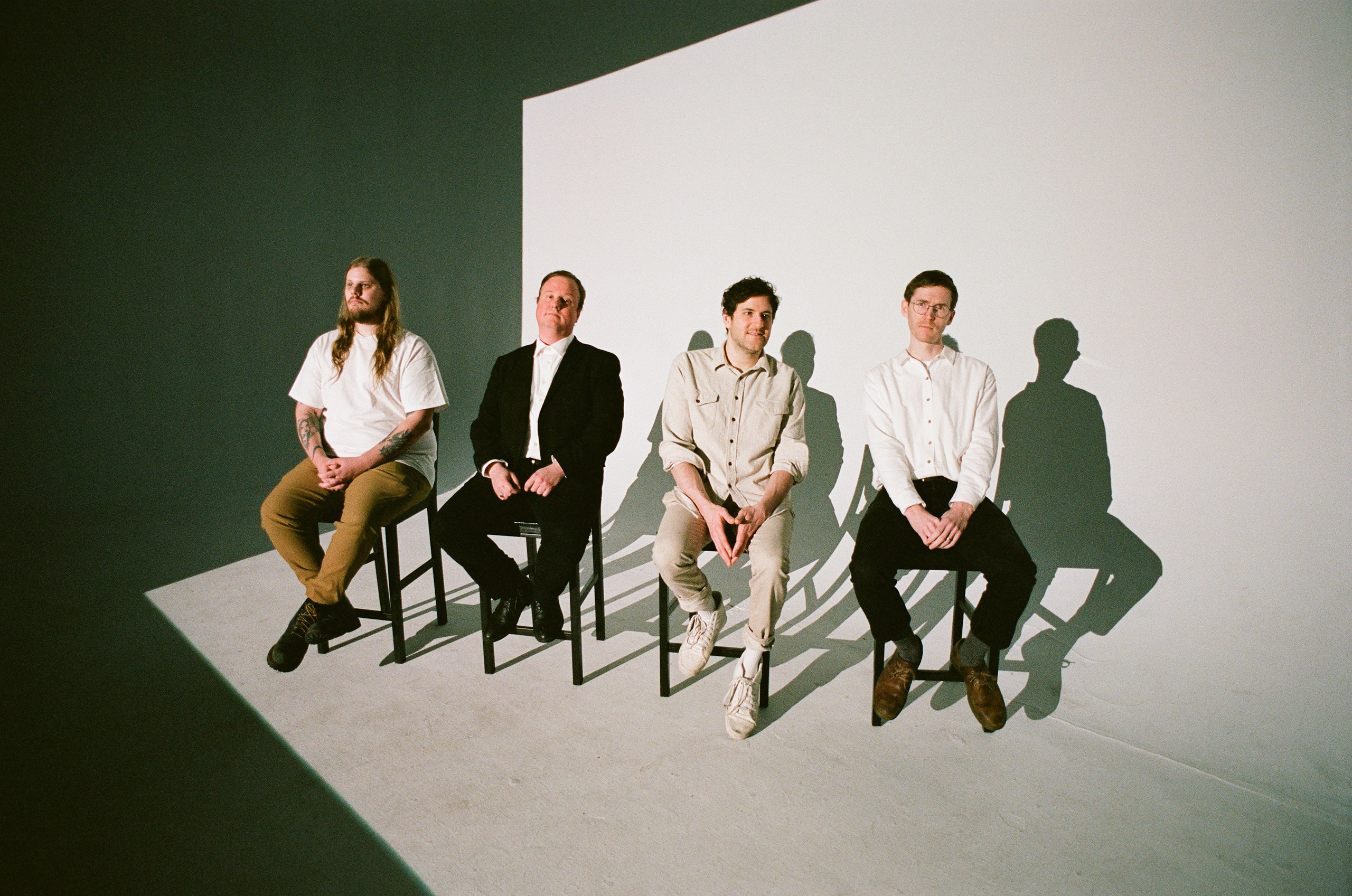 The form band members of Protomartyr sit on a row of chairs in a in a black and white room